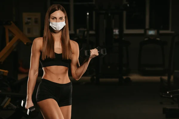 A fit woman in a face mask to avoid the spread of coronavirus is doing bicep curls with dumbbells. A sporty girl in a surgical mask is posing during the arms workout in the gym.