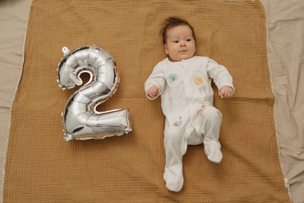 かわいい赤ちゃんは 2の形をした銀の箔風船の近くのムスリン毛布に横たわっていて 足を上げています ワンピースの服を着た女の子が2ヶ月を祝っている — ストック写真