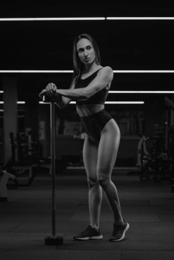 Spor salonundaki kısa bir halterin üzerinde elleriyle poz veren uzun boylu bir kadının fotoğrafı. Kaslı esmer bir kız siyah bluz giyer ve kısa şort giyer..