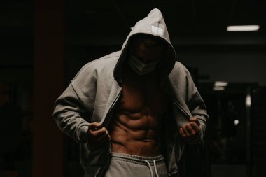 Kapüşonlu ve yüz maskeli bir vücut geliştirici koronavirüsün yayılmasını önlemek için karın kaslarını göstermek için fermuarlı kapüşonlusunu açıyor. Ameliyat maskeli sporcu bir adam antrenmandan sonra poz veriyor.