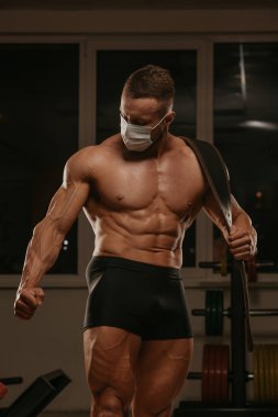 Spor yaptıktan sonra poz veren, tıbbi maske takan üstsüz bir vücutçunun orta boy bir pozu. Cerrahi maskeli kaslı bir adam spor salonunda kaslarını geriyor..