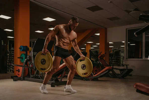 A bodybuilder in shorts is walking through the gym with two rubber plates. A shirtless sporty guy is training alone.