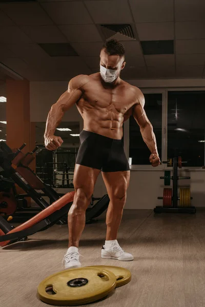 A shirtless bodybuilder in a medical face mask to avoid the spread of coronavirus (COVID-19). A muscular guy in a surgical mask is posing after a workout in a gym.