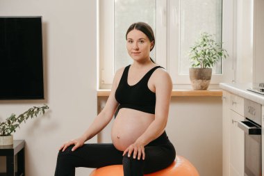 Hamile bir kadının portresi. Turuncu bir egzersiz topunun üzerinde oturuyor ve evindeki mutfağın yakınındaki bir spor salonundan sonra dinleniyor. Sağlıklı hamilelik.