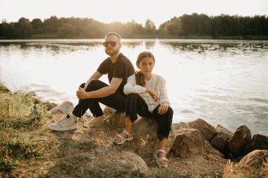 Sakallı ve güneş gözlüklü genç bir baba, akşam göl kıyısında güzel kızıyla birlikte taşların üzerinde poz veriyor. Tatilde olan tek ebeveynli şık bir aile..
