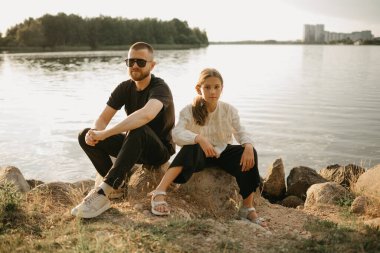 Sakallı ve güneş gözlüklü genç bir baba akşam vakti kızıyla birlikte gölün kıyısında taşların üzerinde oturuyor. Tatilde olan tek ebeveynli aile.