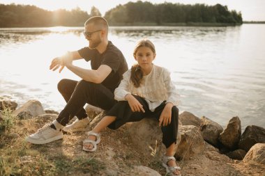 Sakallı ve güneş gözlüklü genç bir baba kızıyla birlikte gölün kıyısında taşların üzerinde oturuyor ve akşam saatlerini kontrol ediyor. Tatilde olan tek ebeveynli şık bir aile..