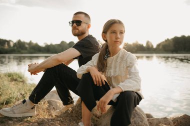 Genç bir kız göl kıyısında sakallı ve güneş gözlüklü babasıyla taşların üzerinde poz veriyor. Günbatımında tatilde olan tek ebeveynli şık bir aile..