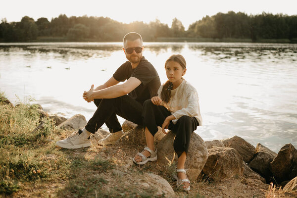 A young father with a beard and sunglasses is posing on stones with his pretty daughter on the coast of the lake. Single-parent stylish family on vacation on the sunset.