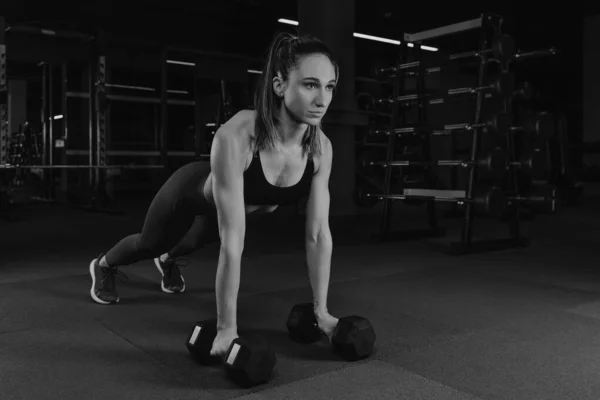 筋肉質の女性はジムでダンベルでプッシュアップされます タンクトップとスポーティーなレギンスのフィット女の子は胸のトレーニングをしています — ストック写真