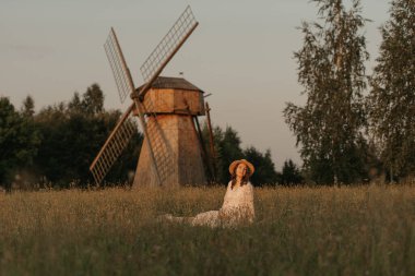 Değirmenin arka planında hamile bir kadın otların üzerinde soğuyor ve akşam çayırda elleriyle karnını tutuyor. Çiftlikte mutlu bir hamilelik. Uzun elbiseli ve hasır şapkalı bir bayan.