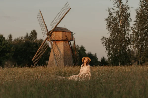 Değirmenin arka planında hamile bir kadın otların üzerinde soğuyor ve akşam çayırda elleriyle karnını tutuyor. Çiftlikte mutlu bir hamilelik. Uzun elbiseli ve hasır şapkalı bir bayan.