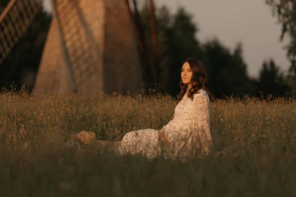 風車の背景には妊婦が草の上に座り 夕方には牧草地で手で腹を抱えている 農場で幸せな妊娠 日没の長いドレスを着た女性 — ストック写真
