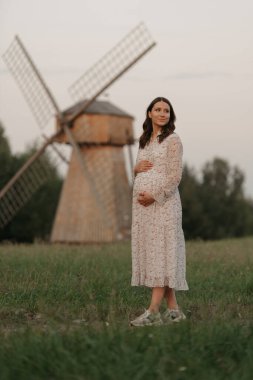 Yel değirmeni arka planında hamile bir kadın akşamleyin çayırdaki çiftlikte elleriyle karnını tutuyor. Mutlu bir hamilelik. Uzun elbiseli bir bayan..