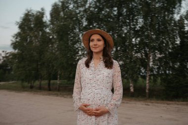 Karnını kum yolunda tutan hamile bir kadının orta uzunlukta bir fotoğrafı. Gün batımında çiftlikte mutlu bir hamilelik. Uzun elbiseli ve şapkalı bir bayan akşam parkta..