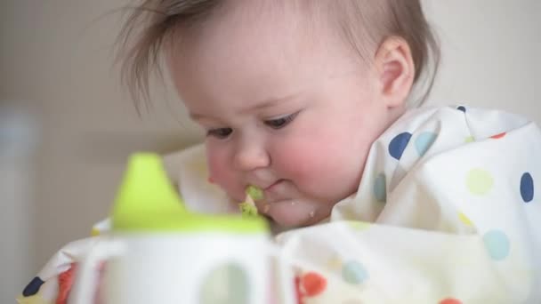 A close-up shot of an infant 7-month girl who is eating broccoli with bare hands — Stockvideo