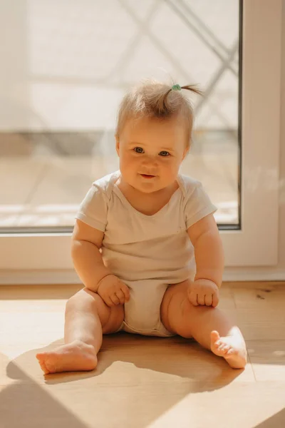 Een meisje van 7 maanden zit bij een balkondeur in een bodysuit thuis. — Stockfoto