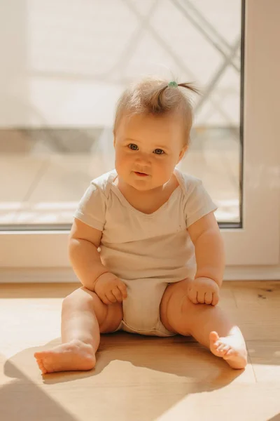 Een vriendelijk meisje van 7 maanden zit bij een balkondeur in een bodysuit thuis — Stockfoto