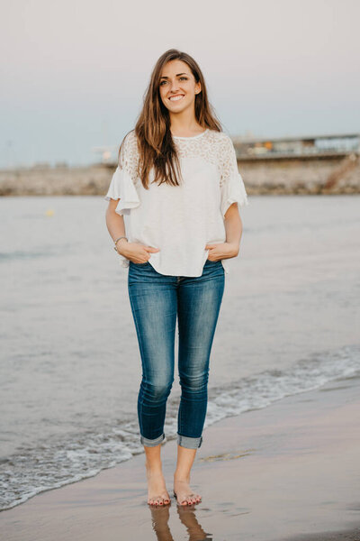 A full-length of a young woman who is standing on a sandy beach near the water and a breakwater at twilight in Spain. A female tourist in the Balearic sea in Valencia.