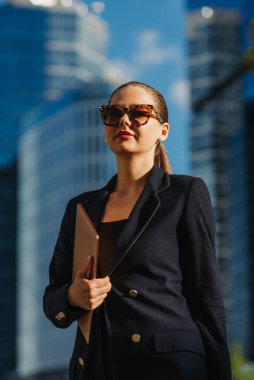 Lacivert ceketli bir bayan emlakçı finans bölgesindeki ofis binaları arasında dizüstü bilgisayarla yürüyor. Bir iş kadını ofisinden yüksek binaların arasında ayrılıyor..