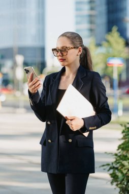 Lacivert ceketli bir bayan emlakçı elinde dizüstü bilgisayar tutuyor ve finans bölgesindeki ofis binaları arasında akıllı telefonunu kullanıyor. Yüksek binaların arasında bir iş kadını..