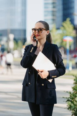 Blazer ceketli bir kadın emlakçı finans bölgesindeki ofis binaları arasında dizüstü bilgisayar tutuyor. Bir iş kadını, yüksek binaların arasında akıllı telefonuyla sohbet ediyor..