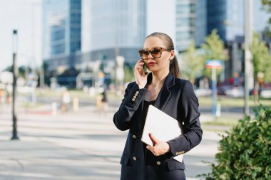 Mavi ceketli bir kadın emlakçı finans bölgesindeki ofis binaları arasında dizüstü bilgisayar tutuyor. Bir iş kadını, yüksek binaların arasında akıllı telefonuyla sohbet ediyor..