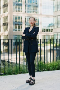 Finans bölgesinde kollarını göğsüne kavuşturmuş spor ceketli bir kadın emlakçı duruyor. Yüksek binaların içinde bir iş kadınının uzun bir fotoğrafı var..