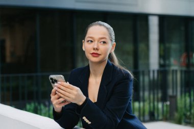 Bir kadın emlakçı, finans bölgesinin modern kentsel alanındaki korkuluklara yaslanırken telefonunu tutuyor. Çağdaş bahçede, yüksek binaların arasında güzel bir iş kadını..