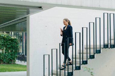 Spor ceketli bir bayanın fotoğrafı, finans bölgesindeki ofis binaları arasında bilgisayarla merdivenlerden iniyor. Çağdaş kentsel alanda güzel bir iş kadını..