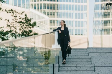 Bir kadın emlakçı, finans bölgesinin merdivenlerinde modern bir kentsel alanda cam korkuluğa yaslanırken bir çanta tutuyor. Gün batımında yüksek binaların arasında bir iş kadını..