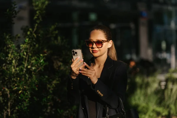 ブレザーの女性のリアクターは 金融街の日没時に現代の庭で携帯電話を持っています 現代の都市空間のかなりのビジネス女性 — ストック写真
