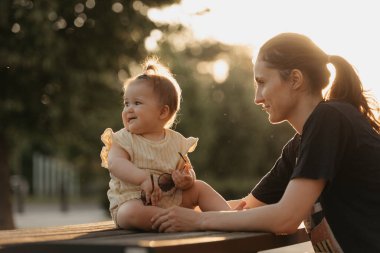 Sarı elbiseli bir bayan çocuk güneş ışınlarında parktaki annesinin yanındaki bankta oturuyor. Anne gün batımında küçük kızının düşmesini engelliyor.