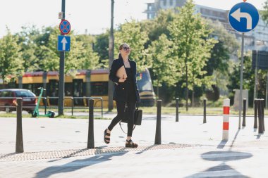 Lacivert ceketli bir kadın emlakçı şehrin merkezinde karşıdan karşıya geçerken dizüstü bilgisayar tutuyor. Bir iş kadını tramvay hattının yakınında geziniyor..