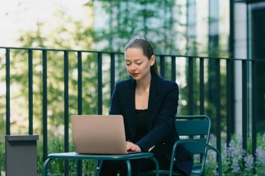 Blazer ceketli bir kadın emlakçı, finans bölgesinde modern bir kentsel alanda dizüstü bilgisayar üzerinde çalışıyor. Çağdaş bahçede bir iş kadını yüksek binaların arasında masada oturuyor.