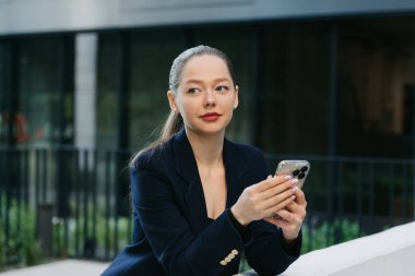 Bir kadın emlakçı, finans bölgesinin modern kentsel alanındaki korkuluklara yaslanırken telefonunu tutuyor. Çağdaş bahçede, yüksek binaların arasında güzel bir iş kadını..