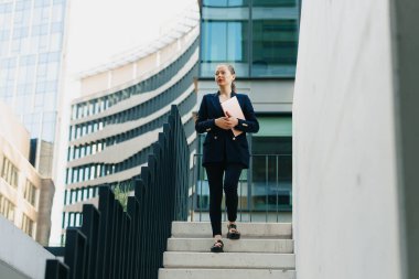 Spor ceketli bir kadın emlakçı, finans bölgesindeki ofis binaları arasında dizüstü bilgisayarla merdivenlerden iniyor. Şehirdeki güzel bir iş kadını yüksek binaların arasında..
