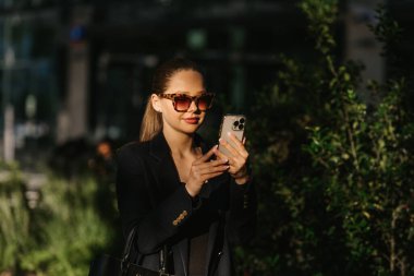 Spor ceketli bir kadın emlakçı, finans bölgesinde günbatımında modern bahçede cep telefonu tutuyor. Çağdaş kentsel alanda güzel bir iş kadını..