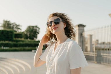 A woman with curly hair in sunglasses is posing in the park at the sunset.