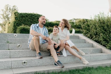 Father, mother, and son are sitting on the steps in the garden of an old European town. Happy family in the evening. Dad is discussing important themes with his smiling family in the park at sunset.