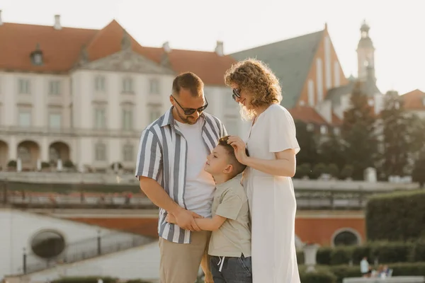 Father, mother and son are hugging in an old European town. Happy family in the evening. Mommy is stroking her little boy\'s head in the background of the palace at sunset.