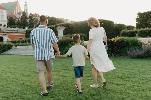 A photo from the back of a happy family is strolling the garden of the palace in an old European town. A smiling father, mother, and son are holding hands and having fun at sunset