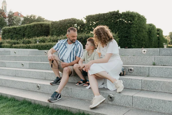 Father, mother, and son are sitting on the steps in the garden of an old European town. Dad is discussing important themes with his smiling family in the park in summer at sunset