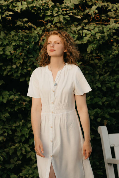 A woman with curly hair is posing in the garden in the evening. A lady in a white dress with green leaves in the background