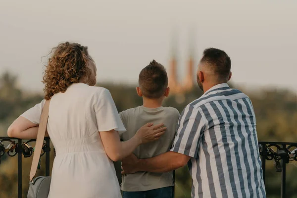 A photo from the back of father, mother, and son who are staring at the old European church. The family of tourists is enjoying the town sight in the evening. Tourists at sunset.