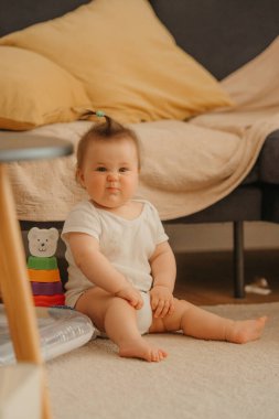 7 aylık bir kız kanepede oturuyor ve evde mayosuyla surat yapıyor. Güneşin altında gülümseyen bir bebek.