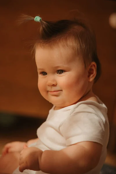 A side portrait of a 7-month girl who is sitting near a dresser in a bodysuit at home. A smiling infant in the sunlight.