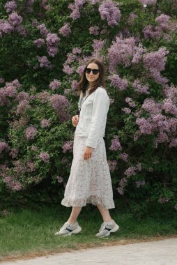 A woman with long hair is staying near the lilac bush in the village green. A lady in black sunglasses and a white dress is posing in the park