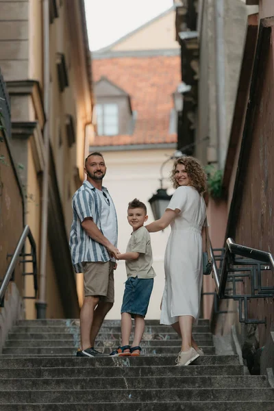 A full-length photo from the back of a family which is turning around while climbing stairs in an old European town. A happy father, mother, and son are holding hands and having fun in the evening.