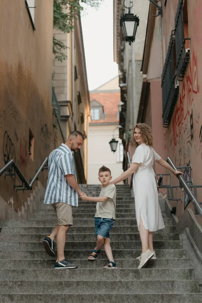 A photo from the back of a family which is turning around while climbing stairs in an old European town. A happy father, mother, and son are holding hands and having fun in the evening.